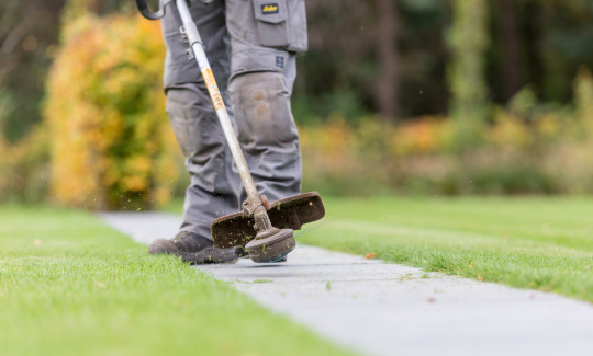 Persoon trimt grasranden langs een tegelpad met een bosmaaier in een gazon.