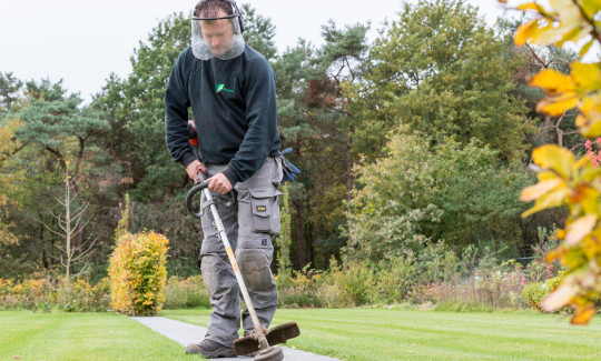 Hovenier met gezichtsbescherming werkt met een bosmaaier aan de grasranden langs een tegelpad in een tuin.