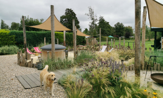 Tuin met natuurlijke beplanting, een houten hot tub, ligstoelen en een golden retriever op een grindpad.