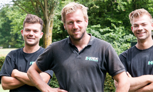 Drie hoveniers in zwarte shirts poseren met gekruiste armen in een groene buitenomgeving.