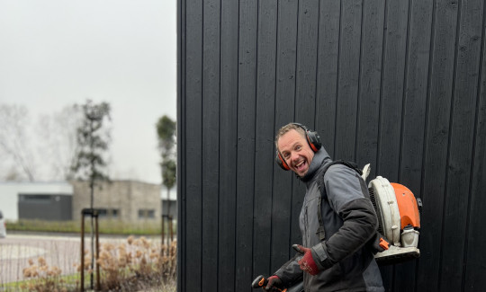 Man met bladblazer op de rug blaast bladeren van een klinkerpad naast een zwart houten gebouw.